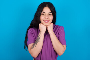 Satisfied young beautiful tattooed girl wearing blue t-shirt standing against blue background touches chin with both hands, smiles pleasantly, rejoices good day with lover