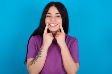 Happy young beautiful tattooed girl wearing blue t-shirt standing against blue background with toothy smile, keeps index fingers near mouth, fingers pointing and forcing cheerful smile