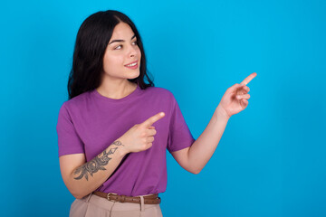 Fototapeta premium young beautiful tattooed girl wearing blue t-shirt standing against blue background smile excited directing fingers look empty space
