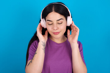 Pleased young beautiful tattooed girl wearing blue t-shirt standing against blue background enjoys listening pleasant melody keeps hands on stereo headphones closes eyes. Spending free time with music