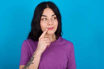 Lovely dreamy young beautiful tattooed girl wearing blue t-shirt standing against blue background keeps finger near lips looks aside copy space.