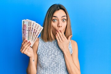 Young caucasian blonde woman holding saudi arabia riyal banknotes covering mouth with hand, shocked...