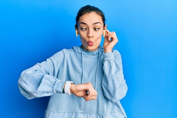 Young caucasian blonde woman using earphones and smart watch making fish face with mouth and squinting eyes, crazy and comical.