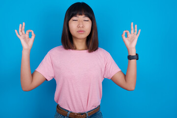 young beautiful asian woman wearing pink t-shirt against blue wall doing yoga, keeping eyes closed, holding fingers in mudra gesture. Meditation, religion and spiritual practices.
