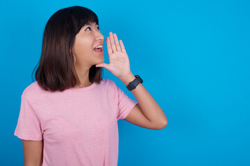 young beautiful asian woman wearing pink t-shirt against blue wall profile view, looking happy and...