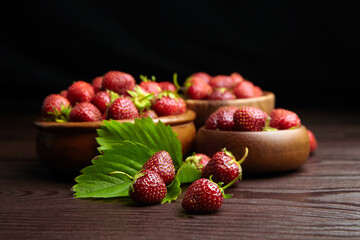 Strawberry red berries in bowls with green leaves on wooden table on black background