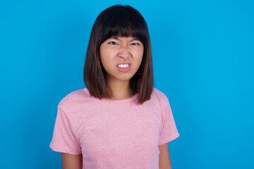 Mad crazy young beautiful asian woman wearing pink t-shirt against blue wall clenches teeth angrily, being annoyed with coming noise. Negative feeling concept.