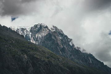 Dark atmospheric surreal landscape with dark rocky mountain top in low clouds in gray cloudy sky. Gray low cloud on high pinnacle. High black rock with snow in low clouds. Surrealist gloomy mountains.