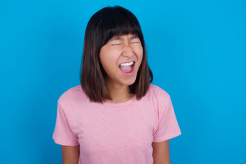 Fototapeta premium Stressful young beautiful asian woman wearing pink t-shirt against blue wall screams in panic, closes eyes in terror, keeps hands on head, finds out terrified news, can't believe it.