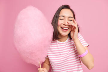 Happy carefree brunette young Asian woman has fun keeps eyes closed holds pink cotton candy enjoys spare free time with friends dressed in casual striped t shirt eats yummy dessert poses indoor