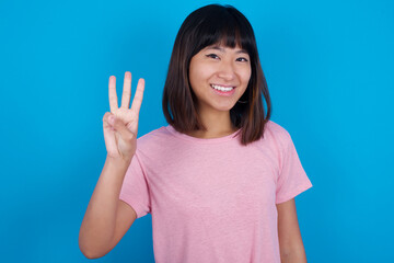 young beautiful asian woman wearing pink t-shirt against blue wall showing and pointing up with fingers number three while smiling confident and happy.