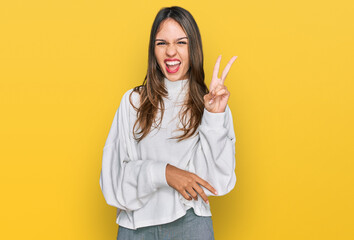 Young brunette woman wearing casual turtleneck sweater smiling with happy face winking at the camera doing victory sign. number two.