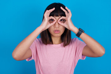 Playful excited young beautiful asian woman wearing pink t-shirt against blue wall showing Ok sign with both hands on eyes, pretending to wear spectacles.