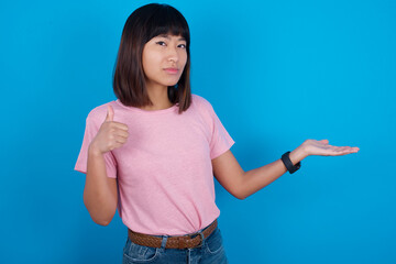 Happy cheerful young beautiful asian woman wearing pink t-shirt against blue wall showing thumb up and pointing with the other hand. I recommend this.