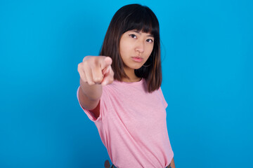 Cheerful young beautiful asian woman wearing purple t-shirt against blue wall indicates happily at you, chooses to compete, has positive expression, makes choice.