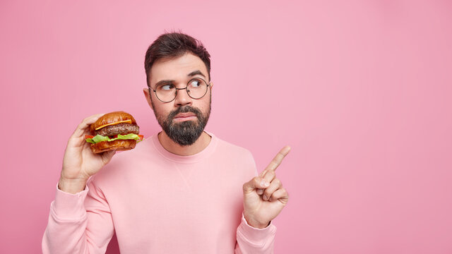 Time for snack. Serious bearded adult man holds delicious burger eats cheat meal points on blank space wears round spectacles casual clothes isolated over pink background. Fast food concept.
