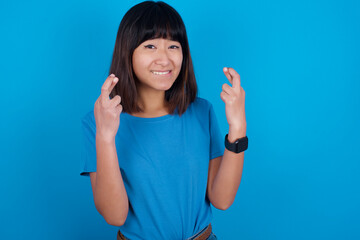 Fototapeta premium Young beautiful asian girl wearing blue t-shirt against blue background holding fingers crossed with worried expression hoping boss didn't noticed mistakes at work.