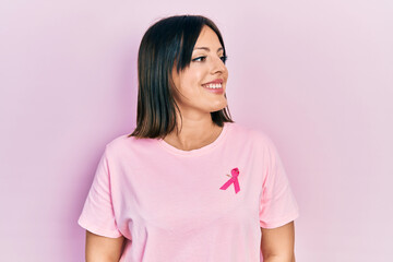 Fototapeta premium Young hispanic woman wearing pink cancer ribbon on t shirt looking away to side with smile on face, natural expression. laughing confident.