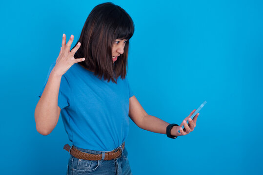 Photo of outraged annoyed Young beautiful asian girl wearing purple t-shirt against blue background holds cell phone, makes call, argues with colleague,  expresses negative emotions. People and anger.