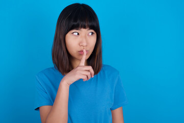 young beautiful asian girl wearing blue t-shirt against blue background makes silence gesture, keeps index finger to lips makes hush sign. Asks not to share secret.
