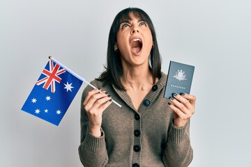 Young hispanic woman holding australian flag and passport angry and mad screaming frustrated and...