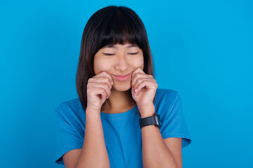 Pleased young beautiful asian girl wearing blue t-shirt against blue background with closed eyes keeps hands near cheeks and smiles tenderly imagines something very pleasant