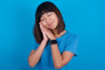 young beautiful asian girl wearing blue t-shirt against blue background leans on pressed palms closes eyes and has pleasant smile dreams about something
