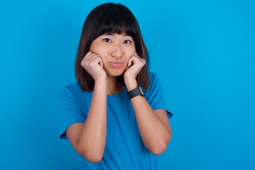 young beautiful asian girl wearing blue t-shirt against blue background with surprised expression keeps hands under chin keeps lips folded makes funny grimace