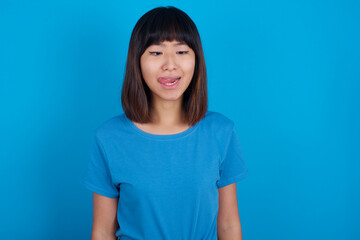 Funny young beautiful asian girl wearing blue t-shirt against blue background makes grimace and crosses eyes plays fool has fun alone sticks out tongue.