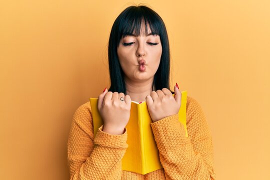 Young hispanic woman holding book making fish face with mouth and squinting eyes, crazy and comical.