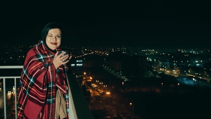 Portrait of a woman on the terrace of a high-rise building on a background of night city and blurred lights. Woman drinking tea or coffee wrapped in a warm blanket.