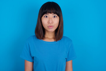 Stunned young beautiful asian girl wearing blue t-shirt against blue background stares reacts on shocking news. Astonished girl holds breath