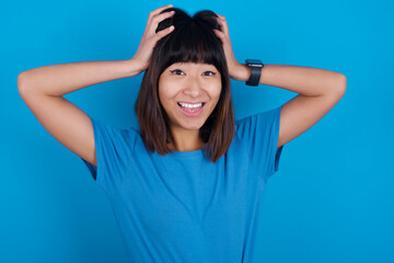 Obraz na płótnie Canvas Cheerful overjoyed young beautiful asian girl wearing blue t-shirt against blue background reacts rising hands over head after receiving great news.