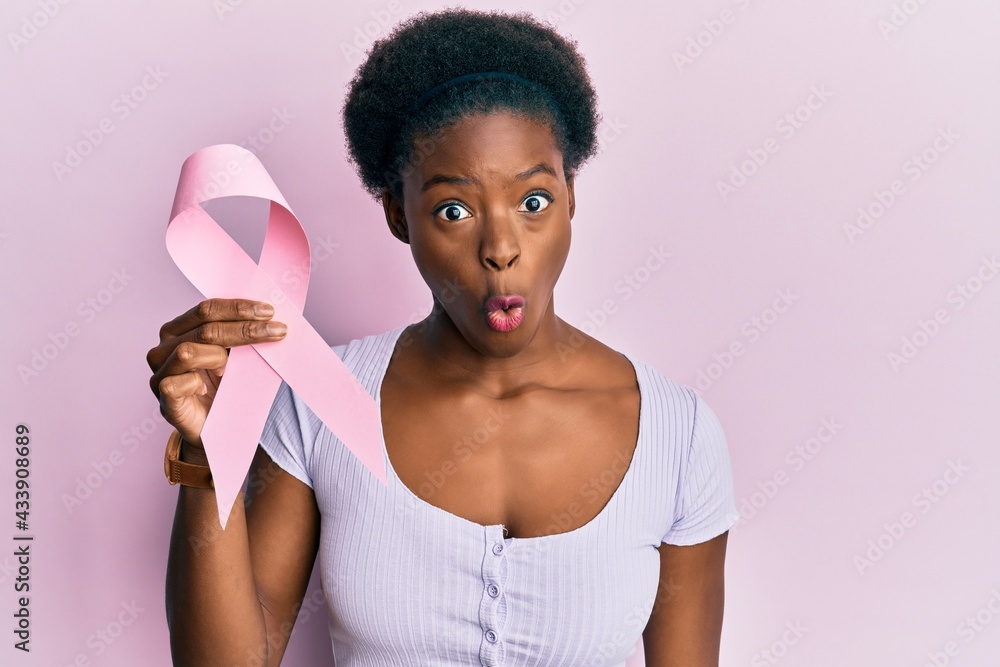 Wall mural Young african american girl holding pink cancer ribbon scared and amazed with open mouth for surprise, disbelief face