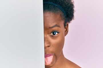 Young african american girl holding blank empty banner over half face sticking tongue out happy with funny expression.