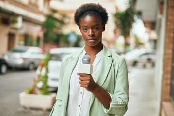 Young african american reporter woman with relaxed expression using microphone at the city