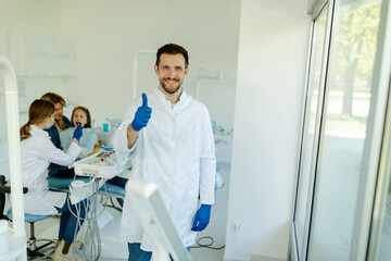 Obraz premium Male dentist standing in dental office, wearing gloves and white coat, showing thumbs up.