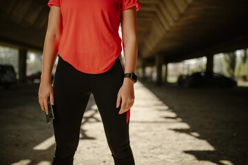 Photo of sporty woman wearing sporty clothes, holding a phone while standing under the bridge.