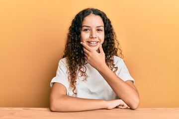 Obraz na płótnie Canvas Teenager hispanic girl wearing casual clothes sitting on the table looking confident at the camera smiling with crossed arms and hand raised on chin. thinking positive.