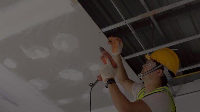  Asian construction worker in safety clothing and work gloves is fastening the drywall ceiling to the metal frame using an electric screwdriver on the ceiling covere