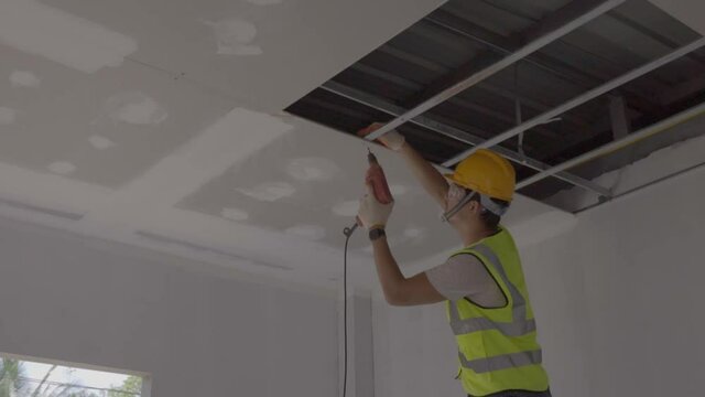  Asian construction worker in safety clothing and work gloves is fastening the drywall ceiling to the metal frame using an electric screwdriver on the ceiling covere