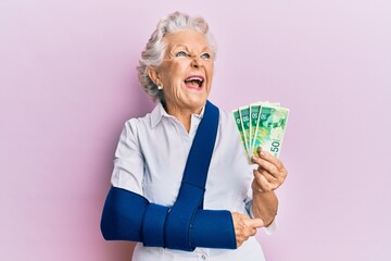 Senior grey-haired woman wearing arm on sling holding israel shekels banknotes angry and mad...