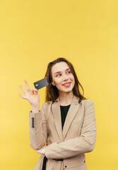 Cute positive lady in a beige jacket poses with a bank card on a yellow background and looks away with a smile on her face.