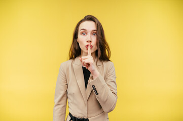 Attractive business woman in a jacket shows a gesture of silence and looks at the camera with a surprised face, isolated on a yellow background.
