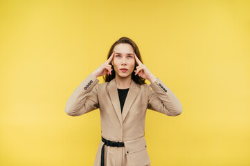 Pretty lady in a suit touches her index fingers to her temple and thinks looking up, isolated on yellow background.