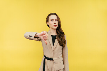 Beautiful woman in a beige suit stands on a yellow background and shows a dislike gesture with a...