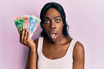 Young african american woman holding south african rand banknotes scared and amazed with open mouth for surprise, disbelief face