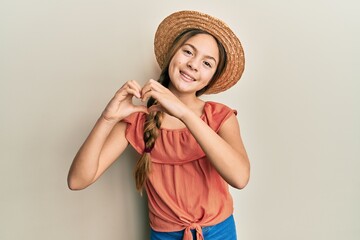 Beautiful brunette little girl wearing summer hat smiling in love showing heart symbol and shape with hands. romantic concept.