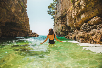 young adult woman in black swimsuit walking into sea between rocks cliff
