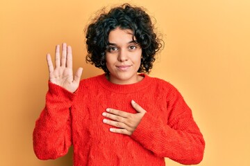 Young hispanic woman with curly hair wearing casual winter sweater swearing with hand on chest and...